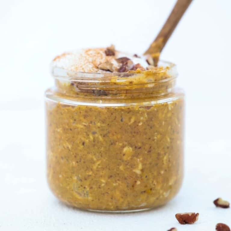 Pumpkin Spice Overnight Oats topped with whipped cream and chopped pecans in a jar with a small wooden spoon.