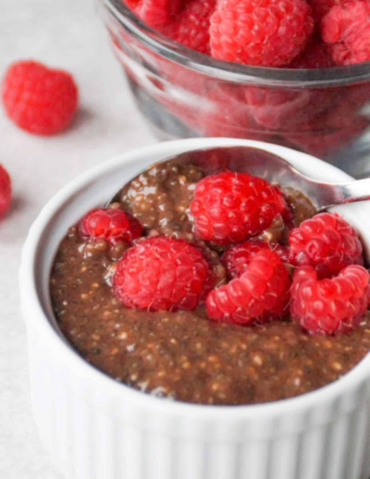 Chocolate chia pudding in a small white ramekin with a spoon and topped with fresh raspberries. One small glass bowl with fresh raspberries is behind the white ramekin.