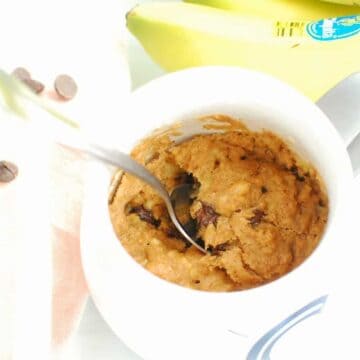 Banana Mug Cake in a white mug with a spoon. One bunch of bananas and a few scattered chocolate chips are in the background.