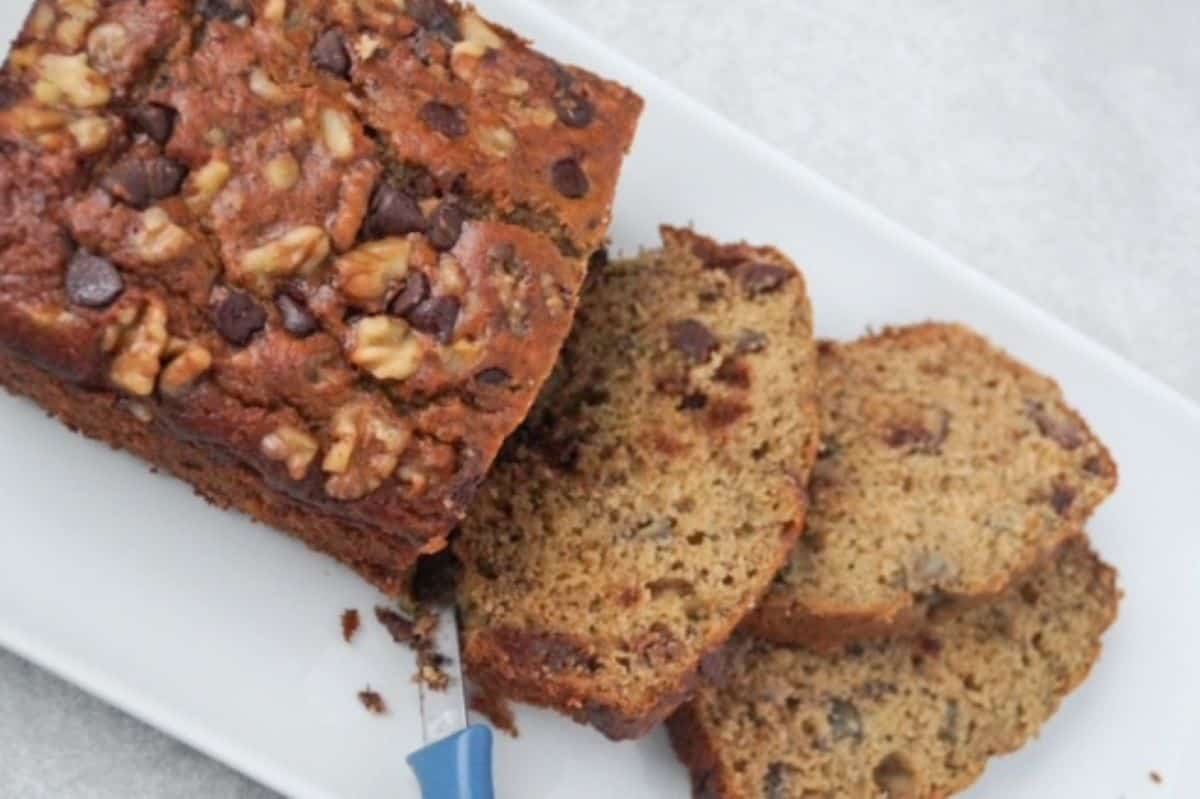 One loaf of banana bread with three slices to the right of the unsliced portion on a white rectangle plate. A paring knife with a blue  handle is shown slicing the piece closest to the rest of the unsliced loaf.