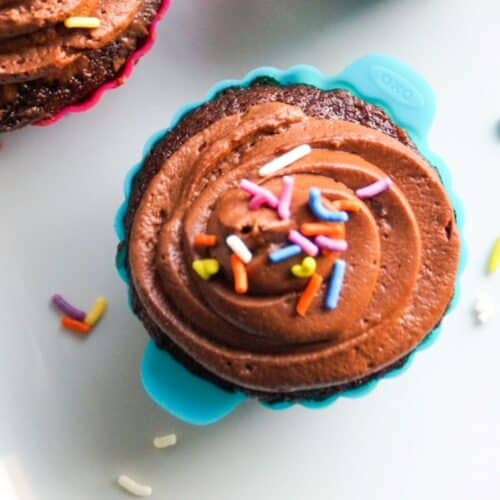 Top view of one chocolate cupcake in a turquoise silicone baking cup, topped with chocolate frosting and sprinkles. The corner another cupcake and a few sprinkles are around the cupcake.