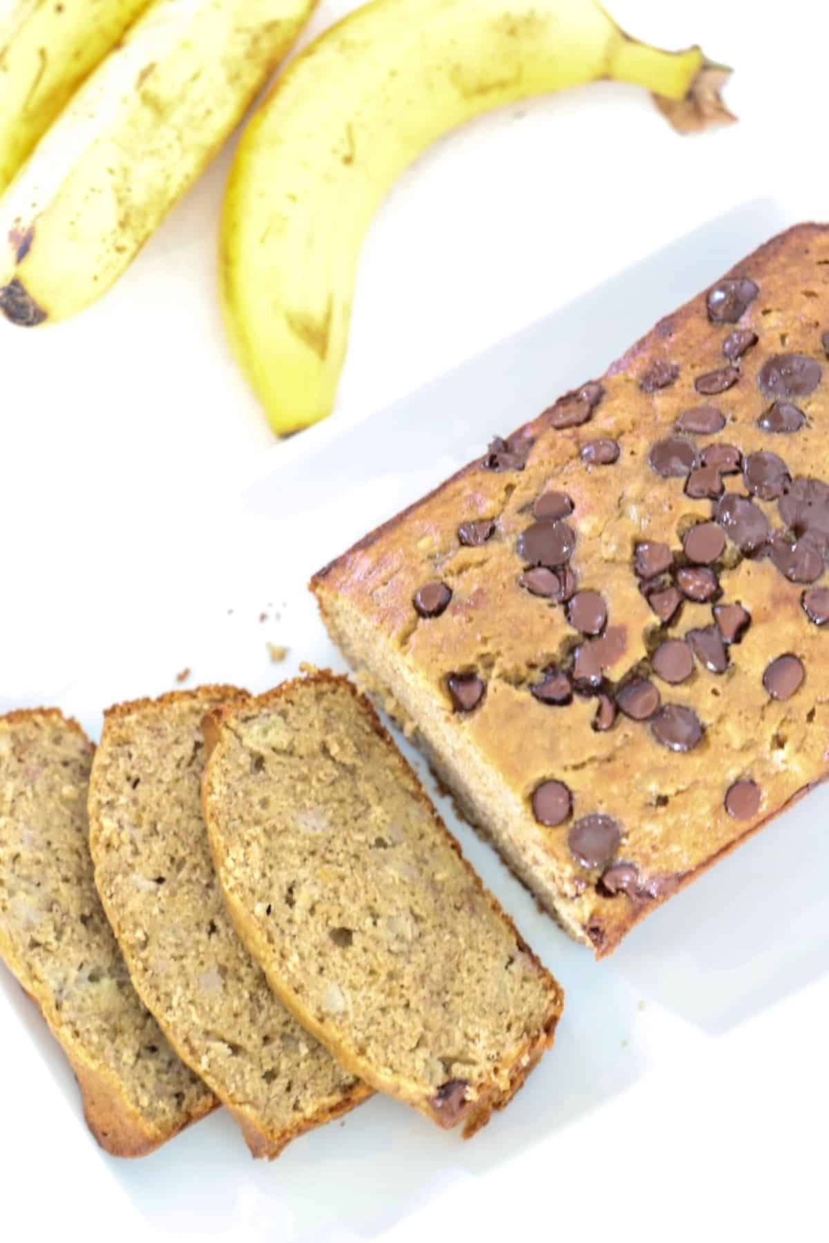 A loaf of banana bread partially sliced on a white platter with bananas in the background.