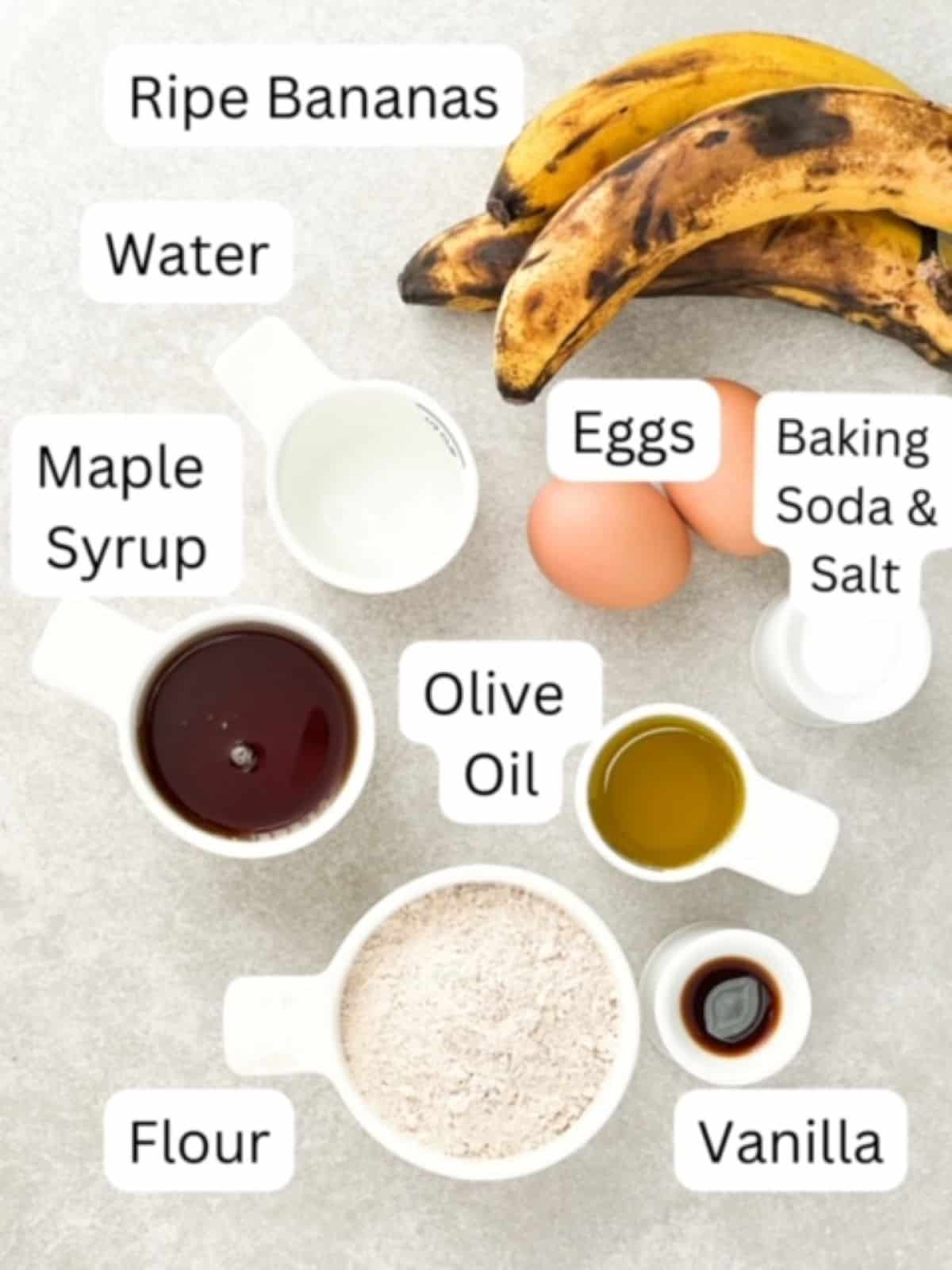 Dairy-Free Banana Bread Ingredients labeled. One bunch of ripe bananas, water, maple syrup, flour, and olive oil in separate white measuring cups, vanilla in a small white prep container, baking soda and salt in a small white prep container, and two brown eggs.