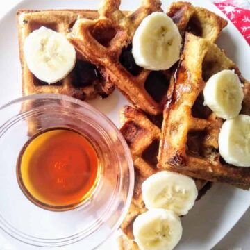 Four quarters of a round waffle overlapping each other on a white plate and topped with banana slices. A small glass bowl with maple syrup is on the plate next to the waffle pieces.