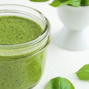 Basil walnut pesto in a glass jar with two basil leaves to the right and a small white egg cup with basil leaves in the background.