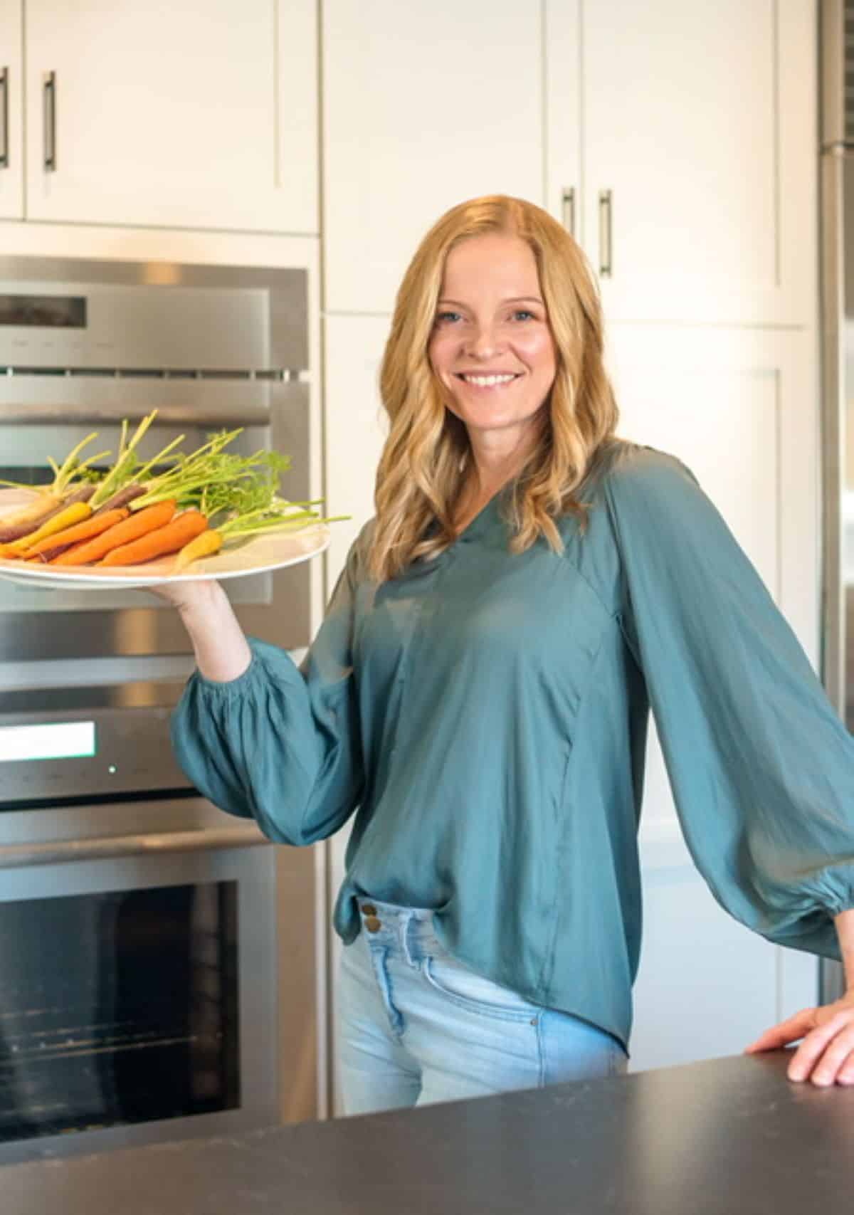 Kristi Ruth RD wearing a green top and jeans holding a white platter with tri-colored carrots.