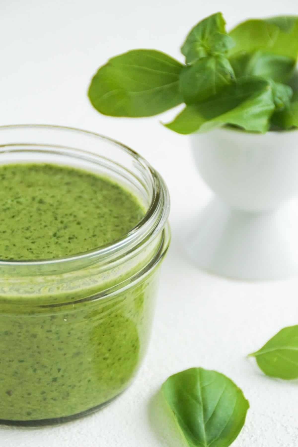 Pesto in a small glass jar with basil leaves to the right of the jar and in a small white dish behind the jar.