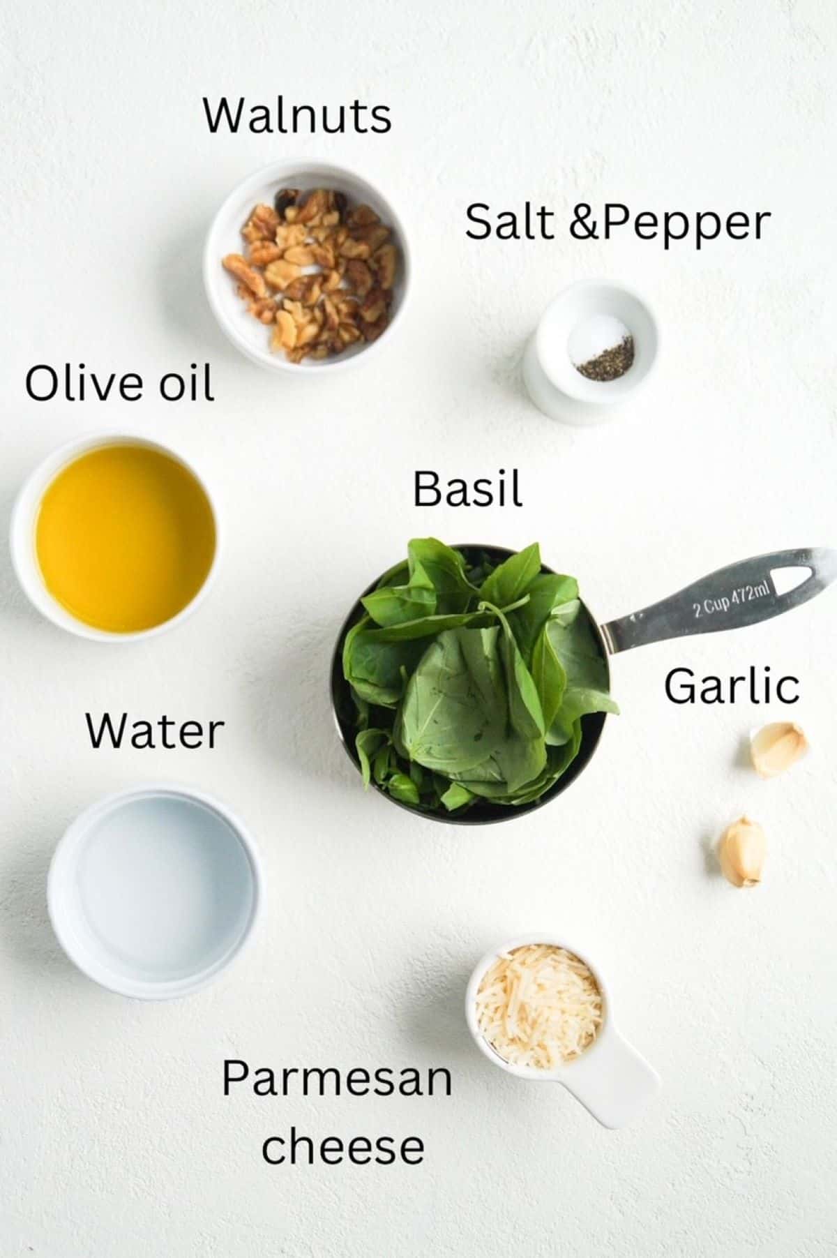 Walnuts, olive oil, water, salt and pepper, and parmesan cheese in individual small white prep bowls, labeled; Fresh basil leaves in a 2-cup measuring cup and two garlic cloves, labeled.