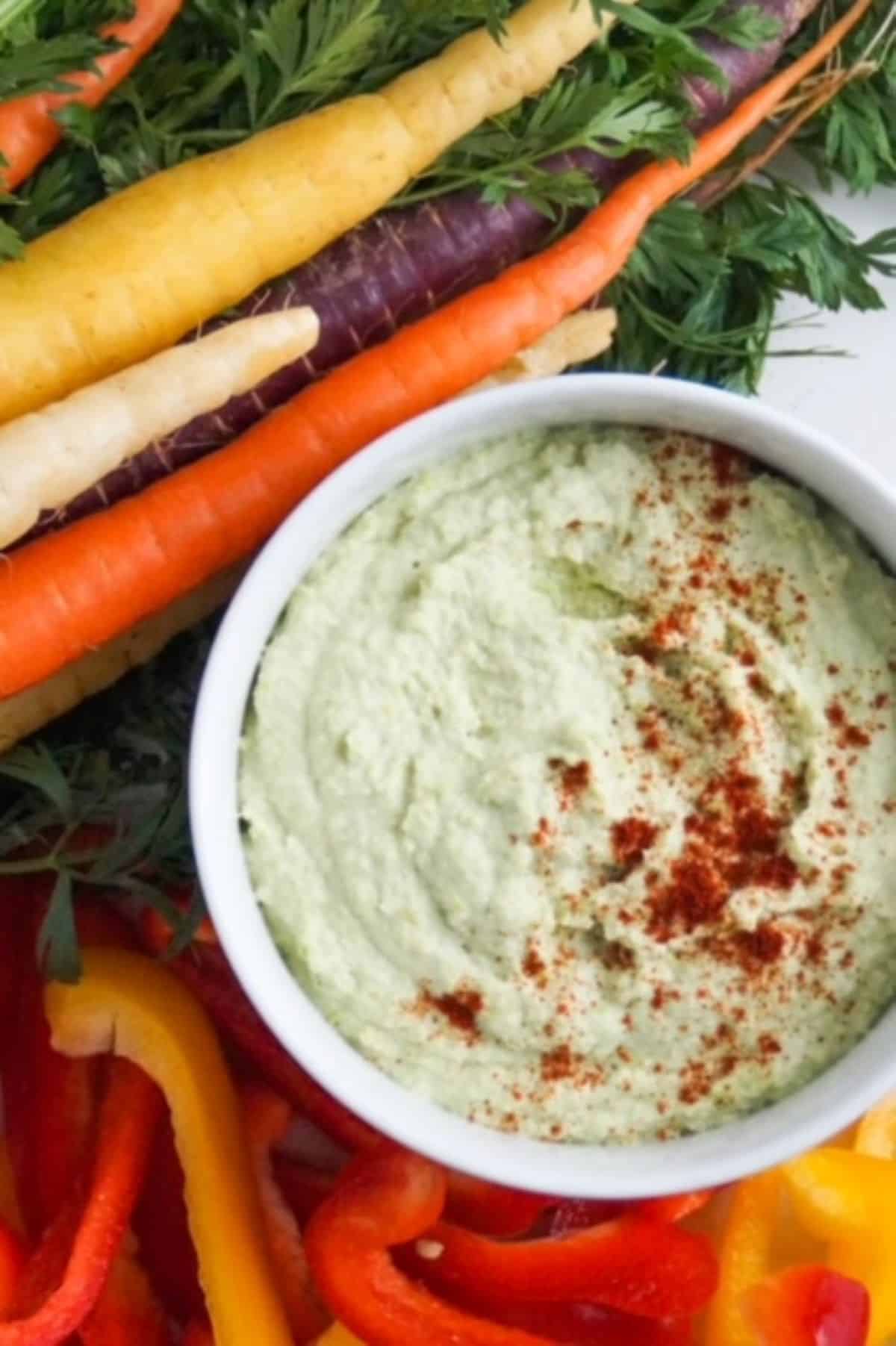 Edamame hummus topped in a white bowl and topped with paprika surrounded by tri-colored carrots and sliced red and yellow bell peppers.