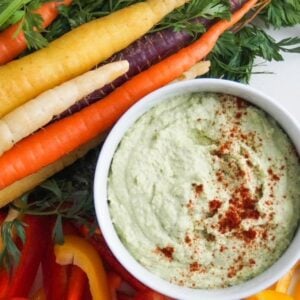 Edamame hummus in a white serving bowl and topped with paprika surrounded by tri-colored carrots and slices of bell pepper.