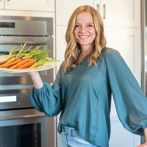 Kristi Ruth RD wearing a green top with jeans and holding a white platter with tri-colored carrots.