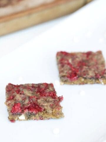 Two Raspberry and White Chocolate Blondies on a white platter with a bar pan of blondies in the background.