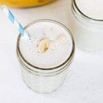 Banana milkshake in a glass topped with banana pieces and served with a blue and white striped straw.