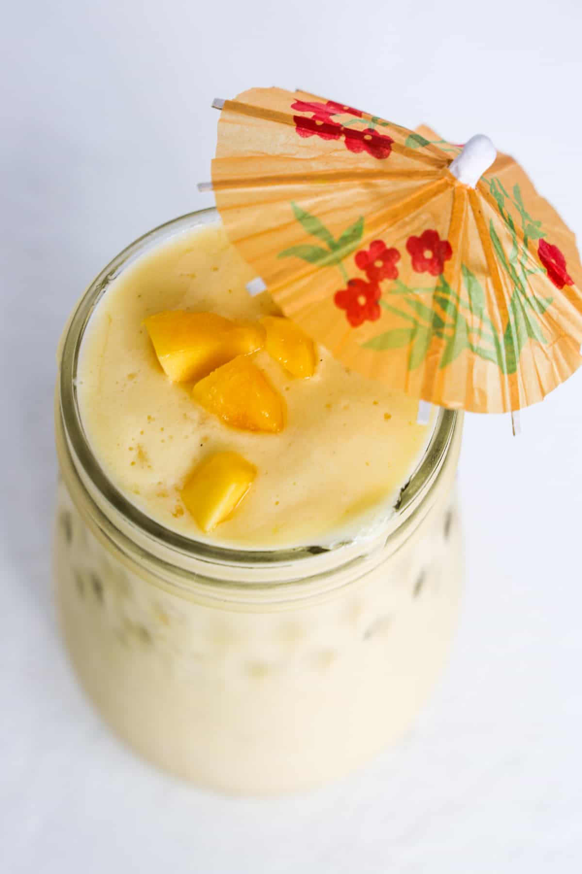 One mango pineapple smoothie in a tall mason jar, topped with pieces of frozen mango and a mini-beverage umbrella.