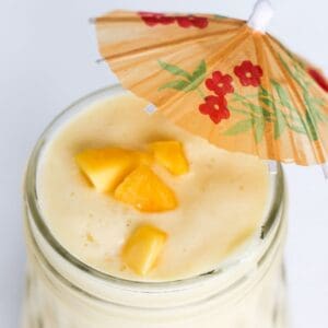 Top view of a mango pineapple smoothie in a glass mason jar and topped with pieces of frozen mango and a mini beverage umbrella.