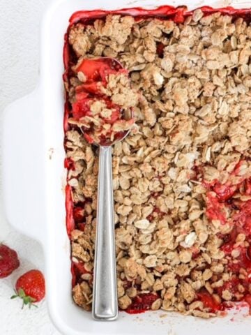 Top view of strawberry crumble in a white square baking dish with one serving of crumble removed and a serving spoon laid on top of the remainder of the crumble.