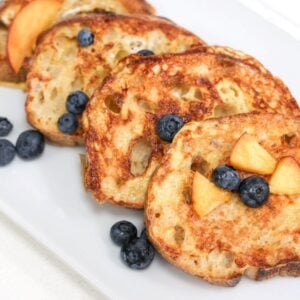 Four slices of sourdough French toast overlapping and topped with peach slices and blueberries.