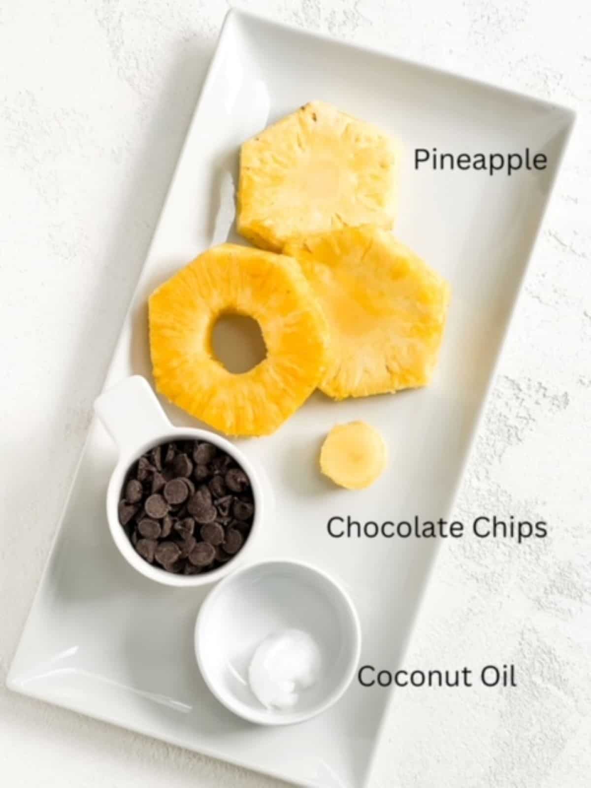 Pineapple rings, chocolate chips, and coconut oil labeled and on a white rectangular plate.