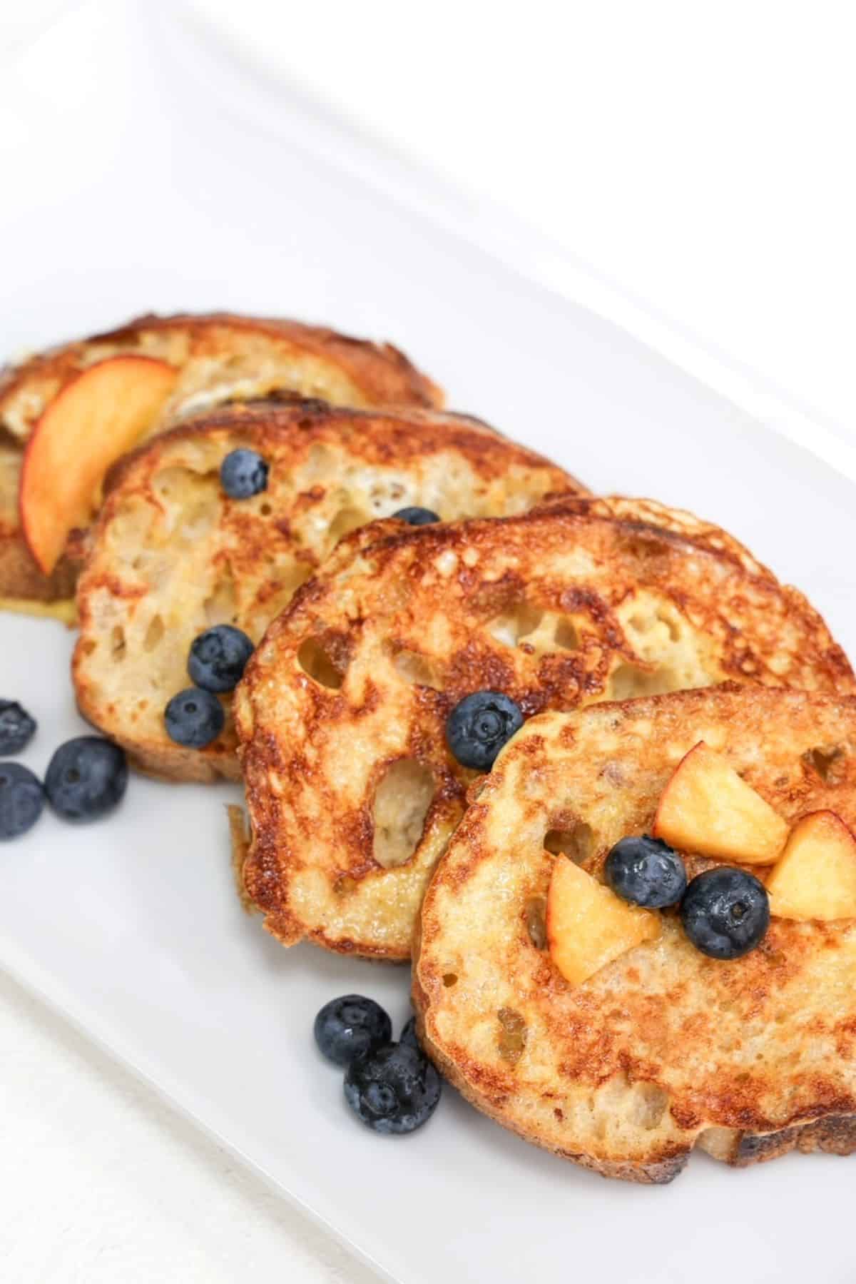 Four slices of sourdough French toast overlapping, topped with blueberries and peach slices.