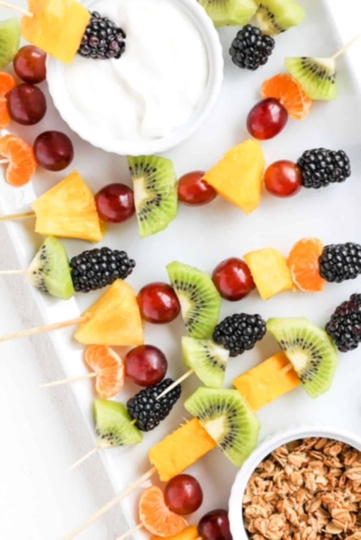 Pieces of fresh fruit on toothpicks and bamboo skewers served on a white plate with granola and yogurt in separate ramekins.