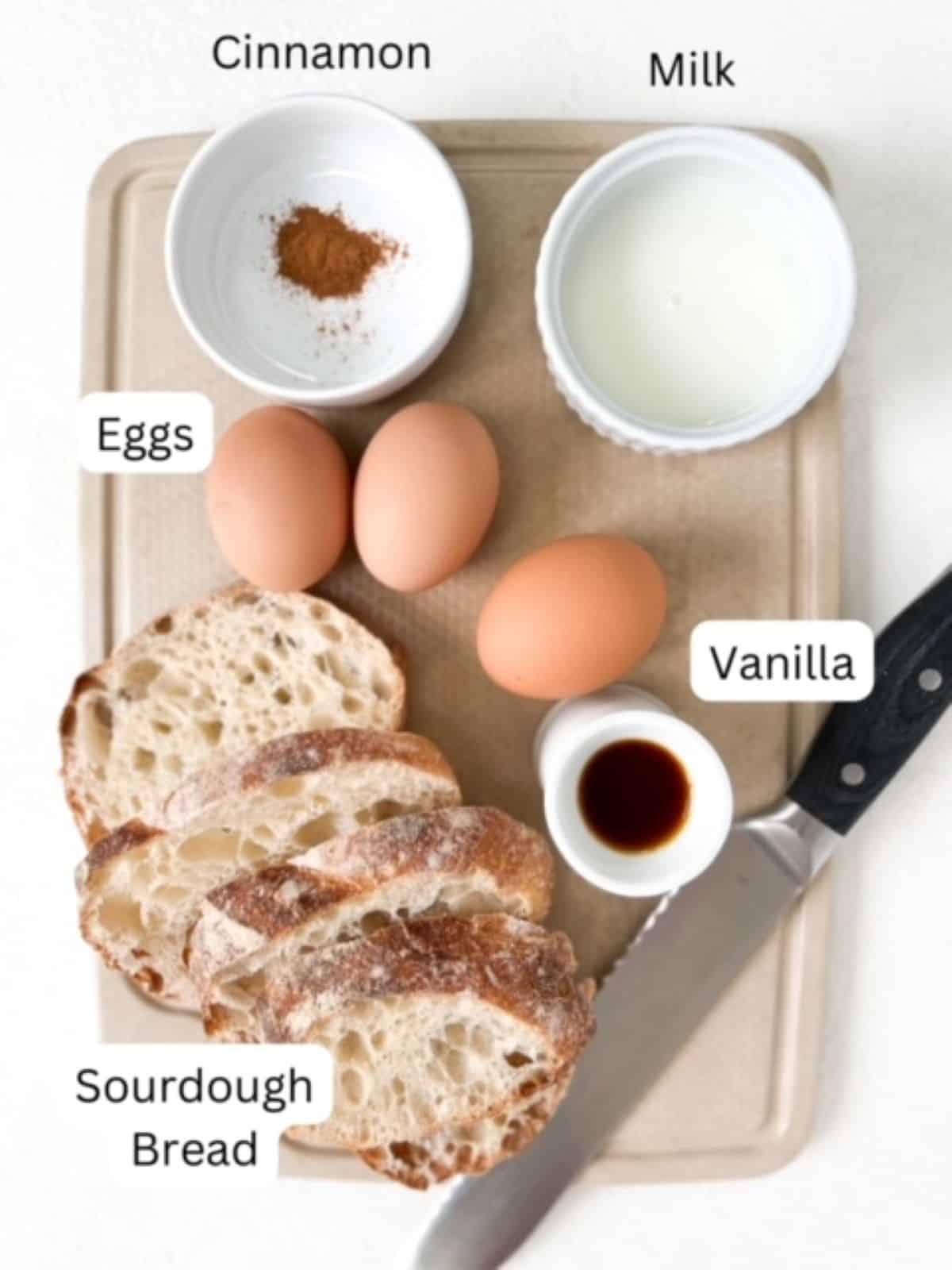 Slices of sourdough bread, milk, eggs, vanilla, cinnamon, and a bread knife on top of a cutting board. All ingredients are labeled.