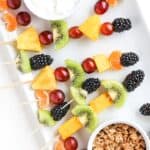 Pieces of fresh fruit on toothpicks and bamboo skewers on a white plate with yogurt and granola in separate ramekins.