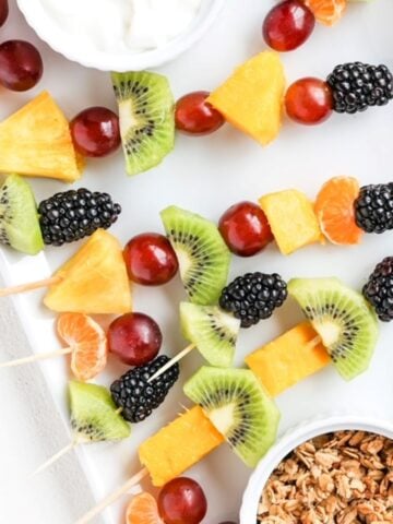 Pieces of fresh fruit on toothpicks and bamboo skewers on a white plate with yogurt and granola in separate ramekins.