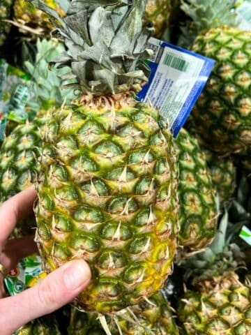 One ripe pineapple with a still on held up over a pile of pineapples.