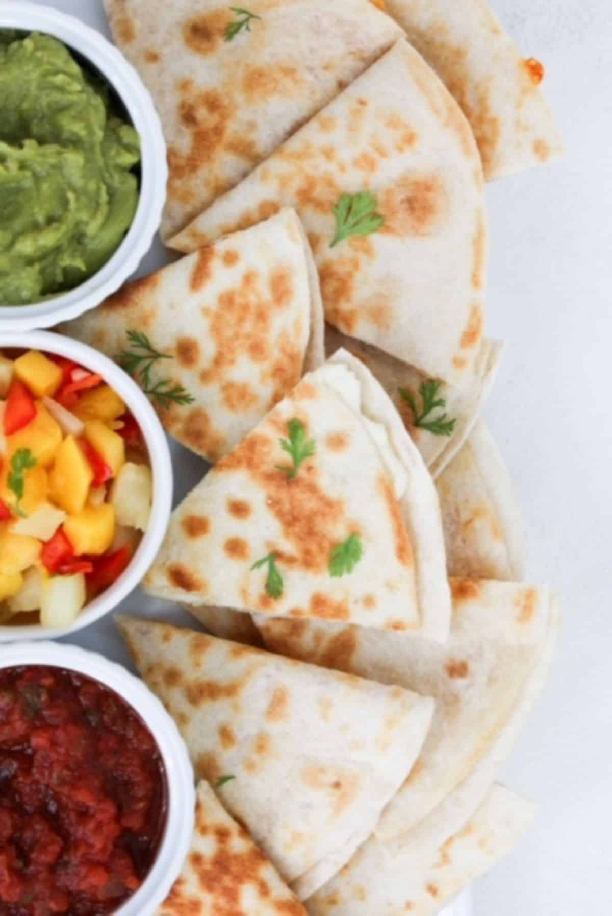 Pieces of cheese quesadillas plated with guacamole, mango pineapple salsa, and tomato salsa in separate white ramekins.