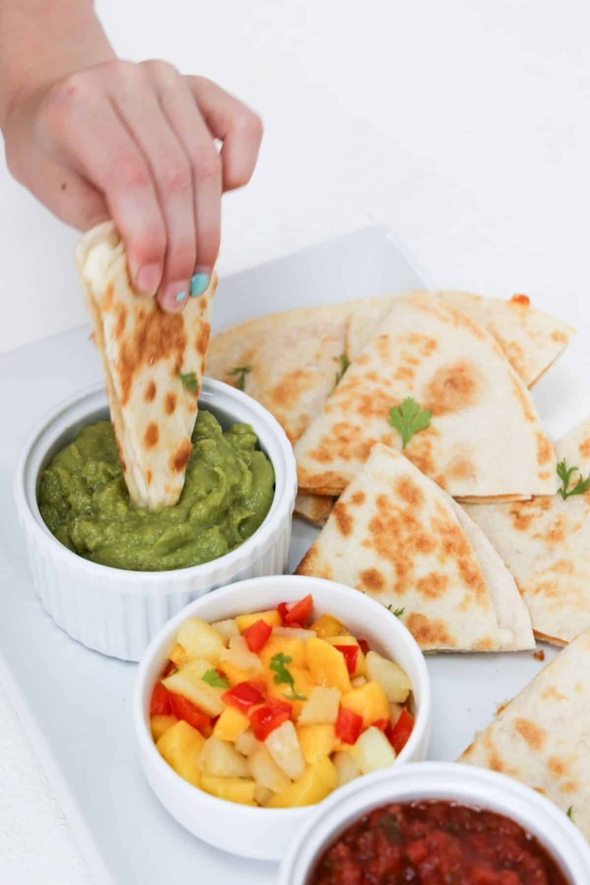 A child's hand dipping one piece of a quesadilla into guacamole plated with more pieces of quesadilla, mango salsa, and salsa.