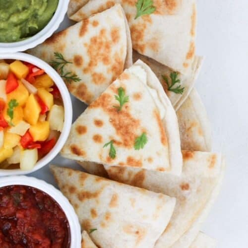Pieces of a cheese quesadilla on a white plate with guacamole, mango salsa, and salsa.