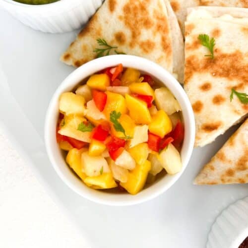 Pineapple mango salsa in a white ramekin with pieces of cheese quesadilla placed around it.