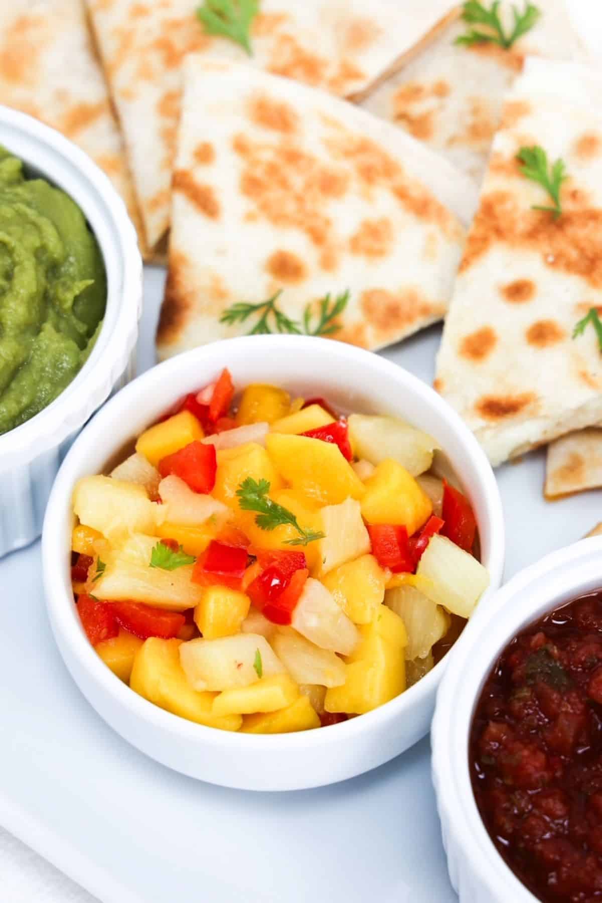Pieces of cheese quesadilla plated with pineapple mango salsa, guacamole, and salsa.