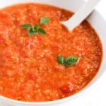 Cherry tomato sauce topped with two small basil leaves and a sprig of fresh oregano served in a white bowl with a white spoon.