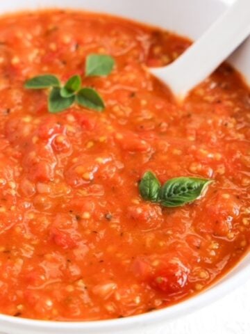 Cherry tomato sauce topped with two small basil leaves and a sprig of fresh oregano served in a white bowl with a white spoon.