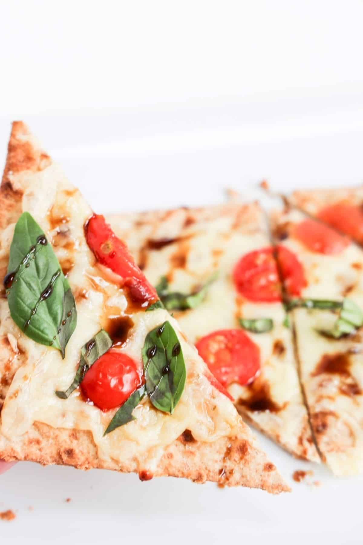 One piece of margherita flatbread held up close over the rest of the flatbread.