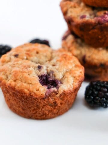 One Blackberry Banana Oatmeal Muffin in the center with a few blackberries and stacked muffins in the background.