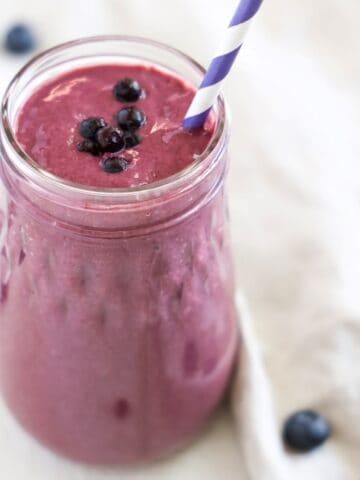 Blueberry Mango Tofu Smoothie with a purple and white striped straw. A linen napken with a few blueberries wraps around part of the base of the smoothie jar.