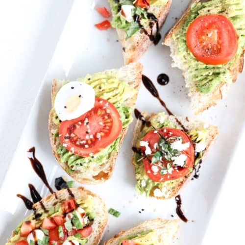 Six pieces of avocado toast topped with tomato and a variety of other topping that differ by piece. Served on a white platter.