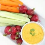 Carrot hummus in a white ramekin with raw veggies on a white rectangular plate.