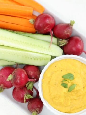 Carrot hummus in a white ramekin with raw veggies on a white rectangular plate.