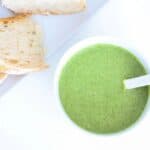 Pesto in a white bowl with a white spoon and a plate of sourdough to the top left of it.