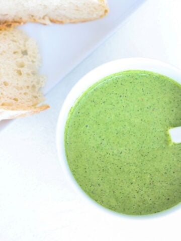 Pesto in a white bowl with a white spoon and a plate of sourdough to the top left of it.