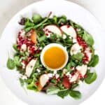 A Christmas holiday salad in a large white serving bowl with a small white bowl with the salad dressing in the center of the salad greens.