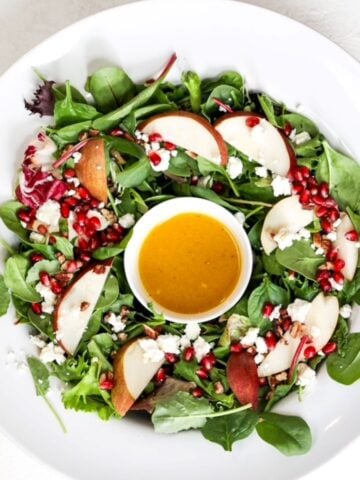 A Christmas holiday salad in a large white serving bowl with a small white bowl with the salad dressing in the center of the salad greens.