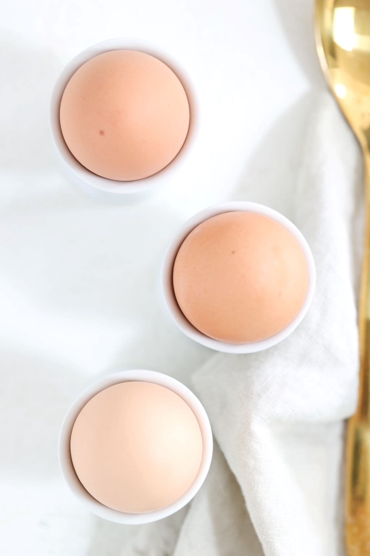 Three brown hard-boiled eggs in white egg cups with a linen napkin and gold spoon to the side.