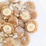 Mini banana pancakes dusted with powdered sugar, topped with sliced banana and almond slices on a white plate.