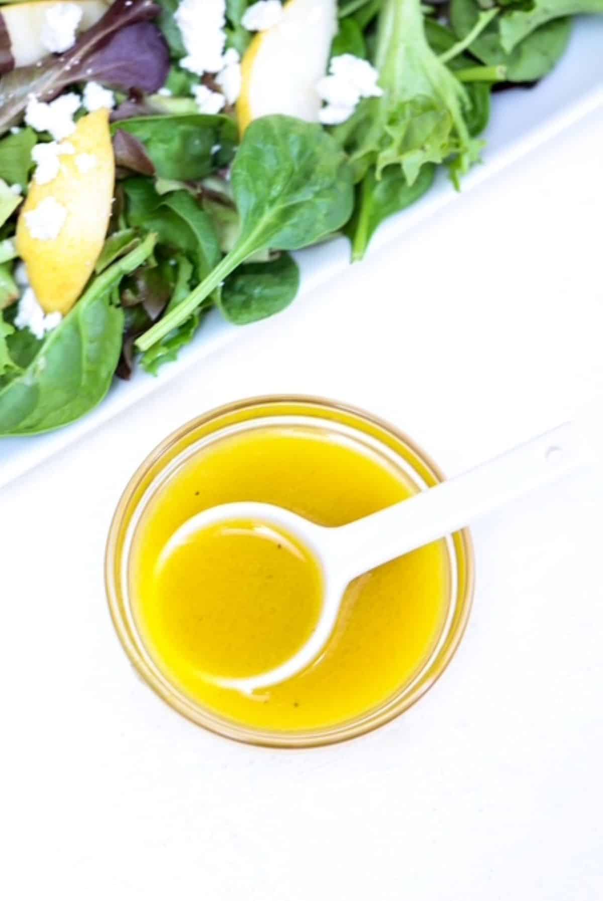Apple cider vinaigrette dressing in a small bowl with a white spoon and a salad to the side.