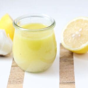 Lemon garlic salad dressing in a glass jar on a white and wooden board with fresh lemon halves and garlic.