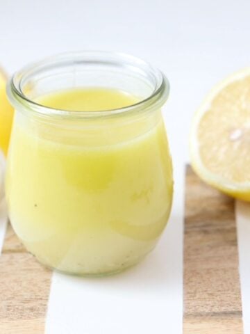Lemon garlic salad dressing in a glass jar on a white and wooden board with fresh lemon halves and garlic.